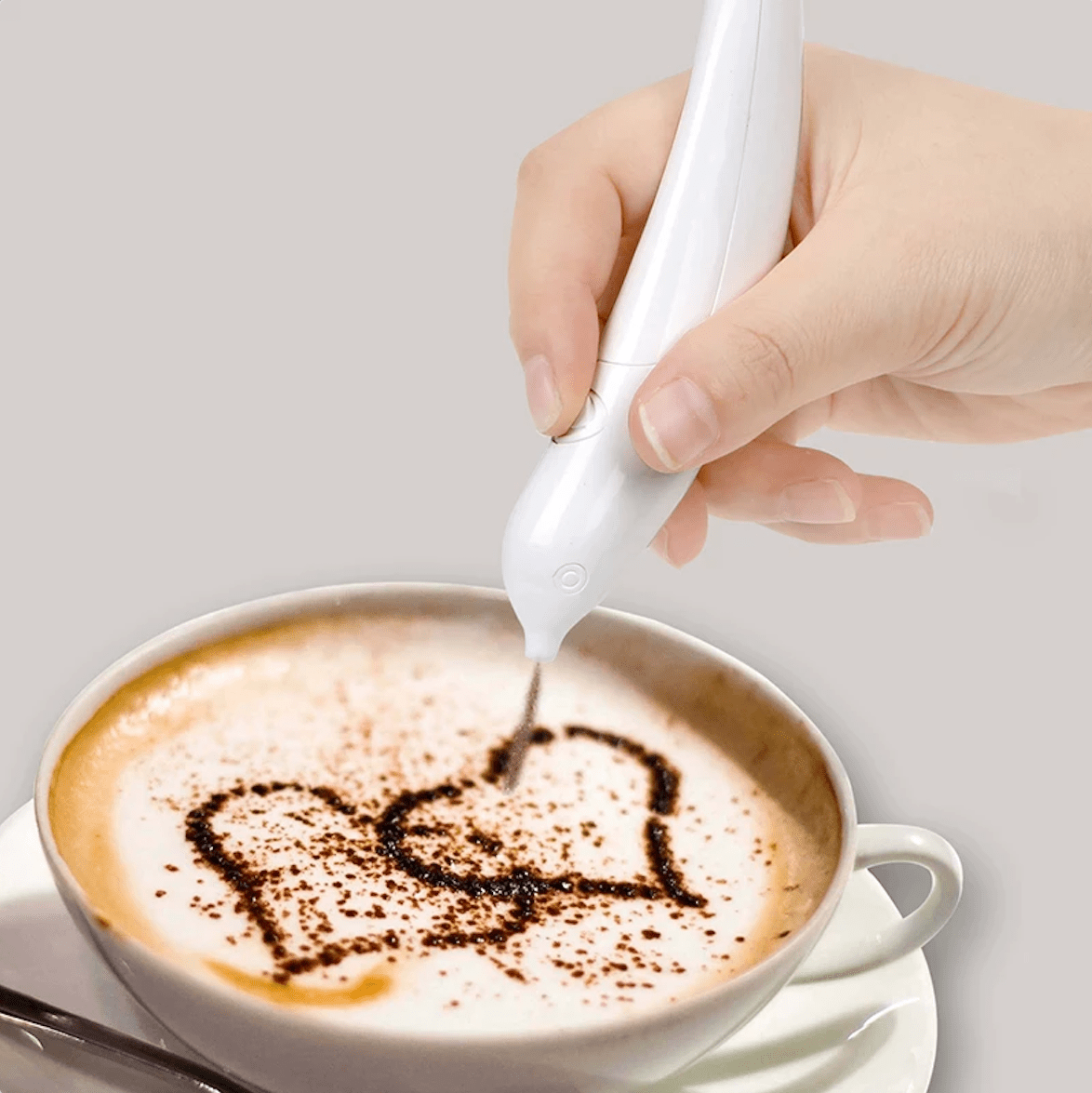 A hand holding a white coffee frothing pen drawing a heart shape on a cup of coffee.
