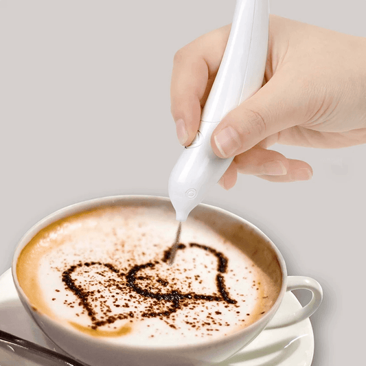 A hand holding a white coffee frothing pen drawing a heart shape on a cup of coffee.